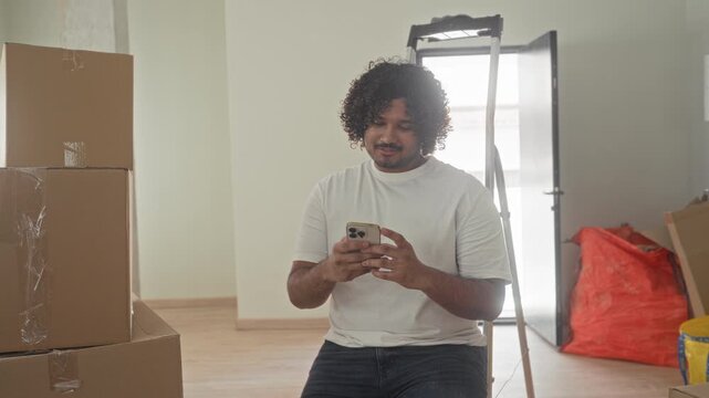 Man holding smartphone with hands checking messages while sitting on ladder amid packed boxes in building; settling contentment.