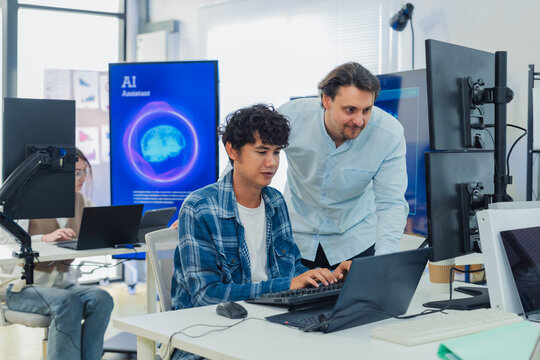 Caucasian mentor pointing at laptop screen while coaching young programmer in modern tech office, perfectly capturing teamwork, IT training, software development, and knowledge transfer.