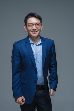 Successful Asian businessman in a blue suit holding a laptop, smiling confidently against a grey studio background, representing professional corporate and technology leadership.