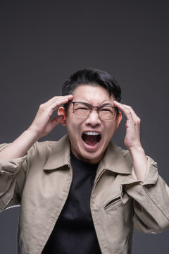 Stressed Asian man with glasses suffering from a severe migraine or tension headache, screaming in pain while touching his temples against a dark grey studio background.