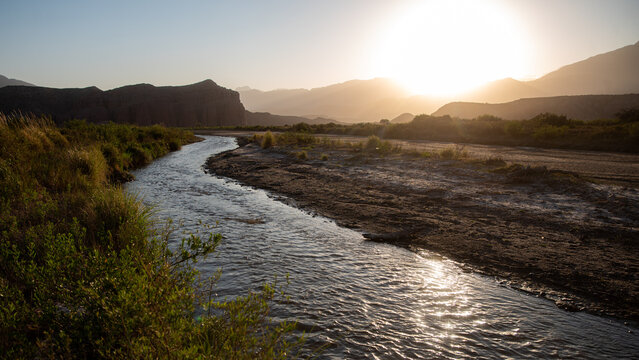 River flowing through a mountain valley at sunset, golden hour light reflecting on water, arid landscape with red rock formations and wild vegetation, peaceful natural scenery.