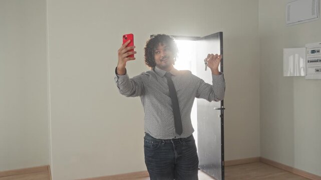 Man in shirt and tie holds smartphone high for a selfie while raising a set of keys in an empty building doorway with sunlight behind him; new home pride.