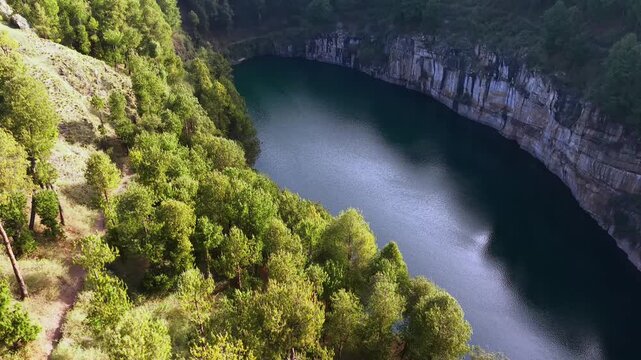 Aerial view shows deep blue crater lake surrounded by lush green forest. Rocky cliffs encircle calm water reflecting sunlight. Path winds along edge offering scenic overlook. Ideal for travel, nature