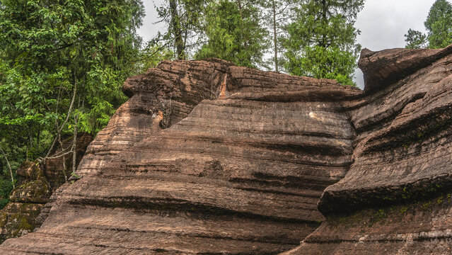Unusual red-brown karst rocks with geological layers on the slopes. Close-up. Green trees against the sky and clouds in the background. China. Red Stone Forest National Geopark. Hunan Province