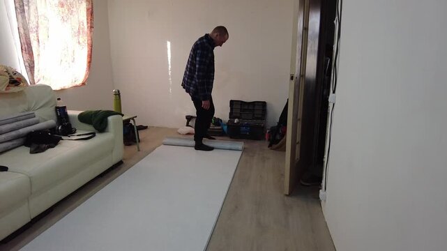 A man prepares a room for wallpapering, rolling out wallpaper on the floor and cutting it to size. The scene takes place in a house with a sofa and light streaming in from a window. Tools and objects 