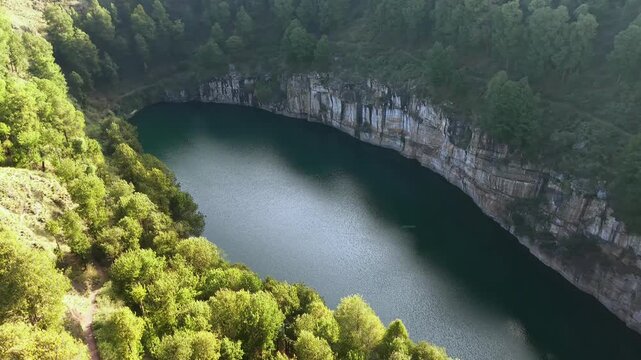 Aerial view reveals a deep, calm crater lake surrounded by lush green trees. Rocky cliffs encircle the dark water, creating a serene natural amphitheater. Shadows stretch across the water