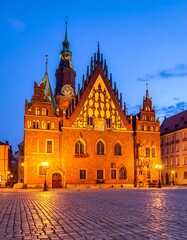 Obraz premium Illuminated facade of an old European city hall at dusk, reflecting lights on the cobblestone square. A towering spire and architectural details are prominent