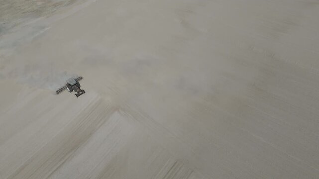 Slow-motion drone footage of a tractor spreading freshly harvested cassava on a large open drying field, representing agricultural processing, food production industrial preparation of cassava starch
