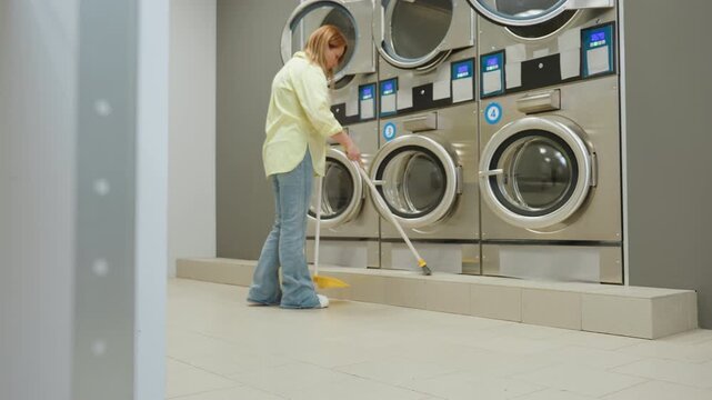 Business owner sweeping floor beside industrial washers in bright laundry room, tidy maintenance routine with broom and dustpan, stainless equipment, tiled surface, shelf and basket in frame