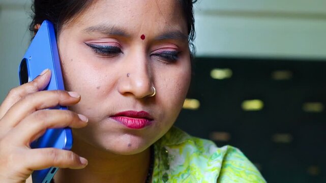 Close-up of a serious Indian woman with a bindi and nose ring talking on a blue smartphone, looking concerned and listening intently.
