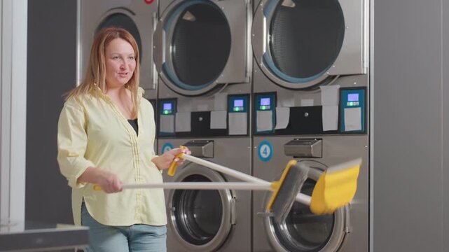 Joyful business woman dances while sweeping near industrial washer in laundromat, holding broom stick and dust pan, cleaning lint spilled from filter drawer on floor during maintenance routine