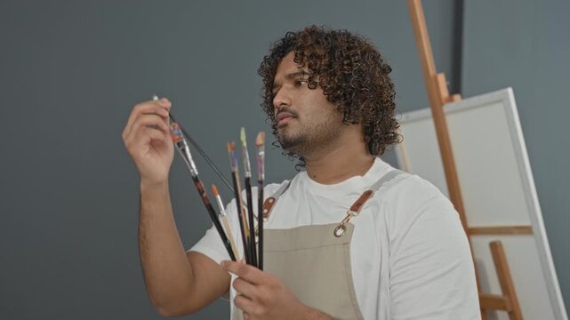 Man painter holding paintbrushes in studio inspecting bristles and choosing brushes near an easel; creative contemplation.