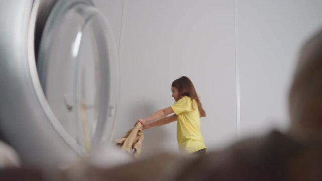 Inside washer view shows happy toddler with blue towel dancing backward, open door left, playful movement in bright laundry room, joy, clean fabrics, round glass rim and handle framing scene