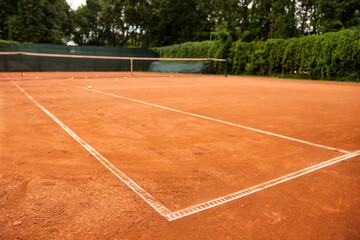 Fototapeta premium A clay court, marked with bright white lines, awaits the next set. Tennis balls scattered across the surface hint at a recent practice. Greenery surrounds the outdoor space.