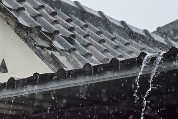 Raindrops fall sharply on a dark, curved tile roof. Water cascades down the gutter in a steady stream. Snowflakes drift softly against a gray sky. The roof shows age, weather, and resilience. © Muhammad