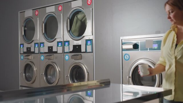 Female entrepreneur carrying cup of tea and notebook walks to table inside laundromat, placing items as industrial washers with digital panels spin clothes in background, showcasing modern service