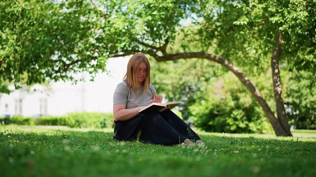 Girl sitting on green grass under leafy tree flipping through book discovering old photo inside, looking at it curiously in warm sunlight, peaceful summer scene filled with nature