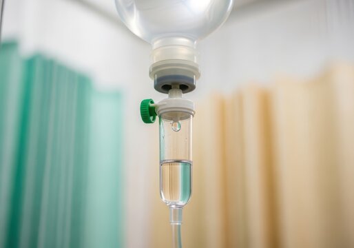 A medical IV drip chamber with a green valve in a hospital room with blurred background curtains