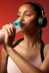 Fototapeta premium Fitness, headphones and woman drinking water in studio for health, body wellness or streaming music. Sports, audio and person with hydration bottle, electrolytes and exercise break on red background