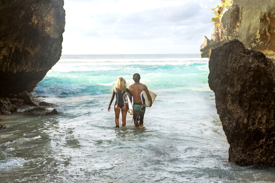 A couple in Bali walks into the ocean with surfboards.