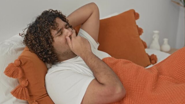 Man holding throat with one hand and clutching chest while lying on bed with orange pillow and textured blanket in studio; illness discomfort.