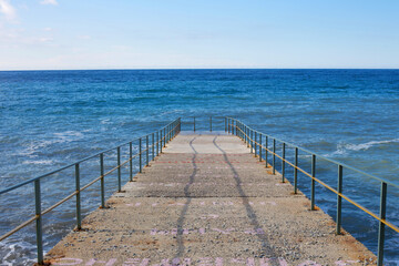 Fototapeta premium Pier leading into blue sea with horizon, minimal seascape. Concrete jetty with railings, calm water and clear sky, symbol of travel, freedom, infinity and peaceful summer vacation