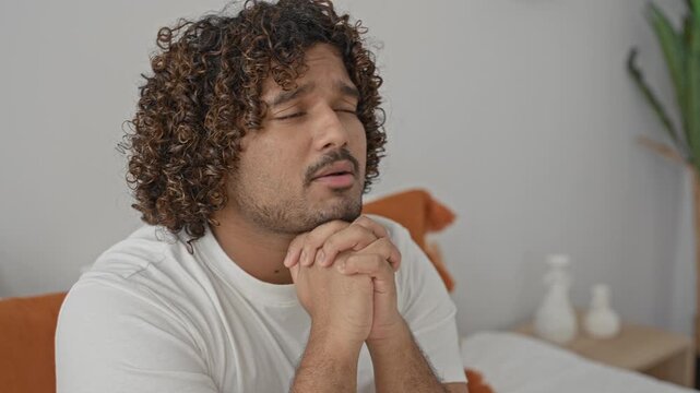 Young indian man on bed in bedroom, white t shirt, eyes closed with hands clasped in prayer and pleading expression, orange pillow and bedside vases visible; quiet devotion.
