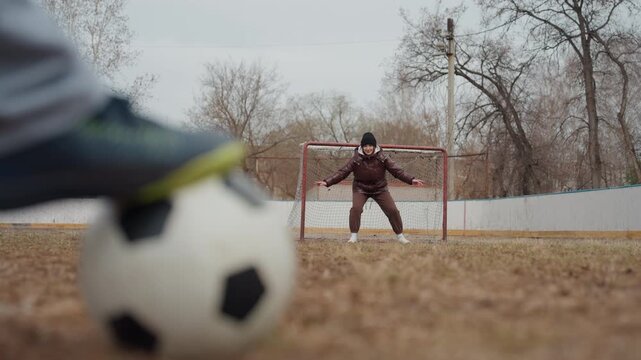 goalkeeper ready stance, ball approaches goalkeeper against overcast sky on vacant sports ground, defensive player in winter gear prepares for action beneath bleak sky on deserted urban football field