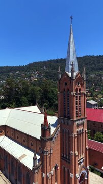 Red brick church with tall spire stands under clear blue sky. Cobblestone paths lead to church entrance. Greenery and hills surround peaceful religious site. Ideal for religious, historical