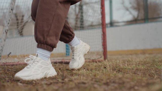 close-up of caucasian feet and sneakers performing agile footwork near goal line, grass and gritty soil underfoot, casual brown sweatpants and white socks visible, rhythm and precision in movement