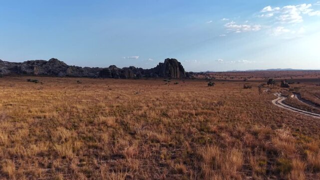 Aerial view of vast, dry savanna with scattered rocky outcrops under clear blue sky. Golden-brown grasses stretch across flat terrain. Scattered trees and shrubs add sparse green accents