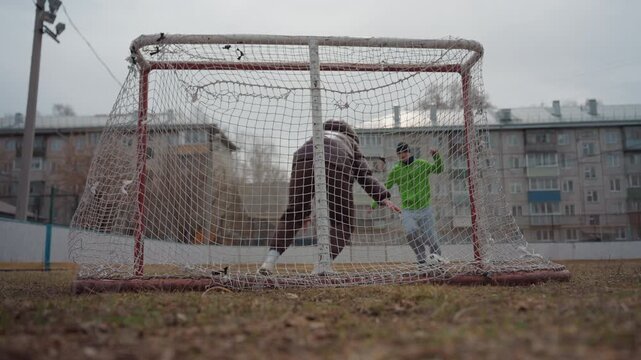 wide goal frame with players and coach, practice drill on urban pitch; mentor guiding young striker in green, routine passes and defensive positioning, overcast community field atmosphere