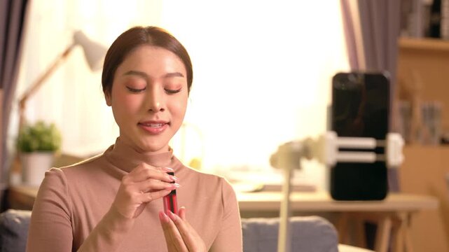 Confident beauty vlogger recording makeup tutorial using smartphone holding lipstick indoors enthusiastic young woman broadcasting engaging live video online from cozy home studio