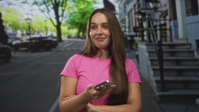 Woman holding smartphone on a street, arms crossed and smiling while tapping screen in pink t shirt; contentment.