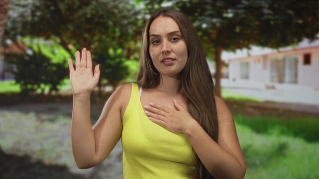 Woman with one hand raised and other hand pressed to bare chest beside a residential building and shaded trees; sincerity.