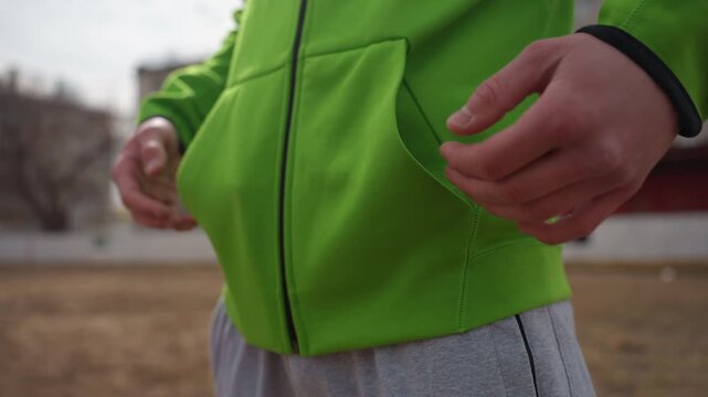 close-up green hoodie pocket outdoor white male. teenager standing on suburban sidewalk with hands near zipper, relaxed posture, overcast sky, fleece texture and muted urban background.