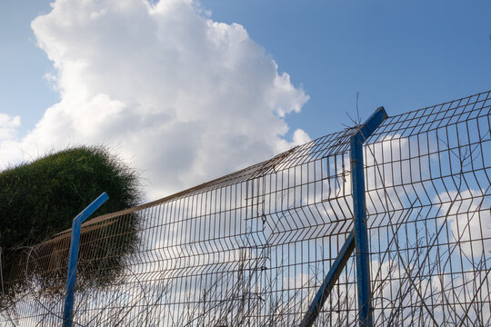 A detailed shot captures a secure wire mesh border fence with blue metal posts and a barb-wire top edge, typical of the demarcation between Israel and Palestinian Authority areas