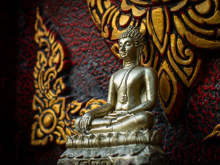Fototapeta premium Silver Buddha Statue Seated in Meditation Against Ornate Red and Gold Thai Temple Wall with Traditional Floral Pattern