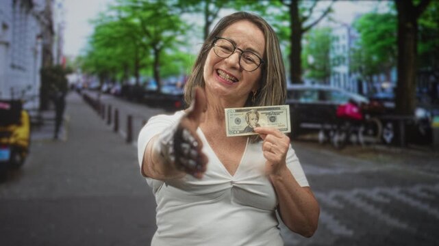 Hispanic senior woman with grey hair and glasses extends right hand for handshake while showing a twenty dollar banknote on an urban street; trust generosity joy.