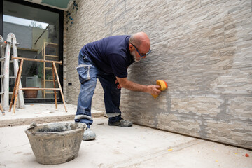 Worker cleaning excess grout from a newly tiled exterior wall with a sponge