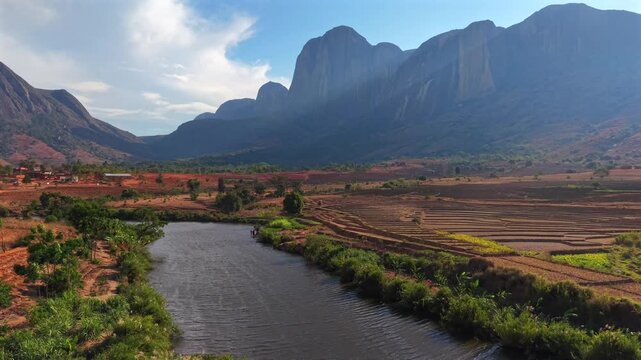 Red soil terraces stretch across valley floor under mountain peaks. River winds through greenery and farmland. Bright blue sky with fluffy clouds above. Scene shows rural agriculture