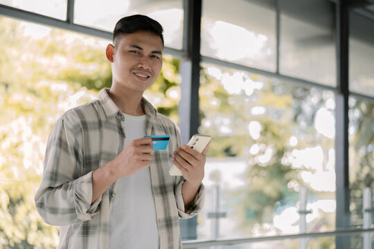 Smiling asian man standing holding blue creadit card and smartphone  in a modern glass hallway.  modern tech. Digital banking. Cashless.