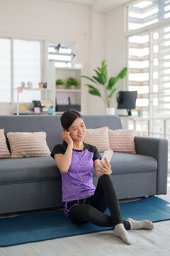 Asian adult woman sportswear enjoy listening music phone app sitting yoga mat home living room modern bright apartment fitness relaxation wellbeing wellness happy lifestyle