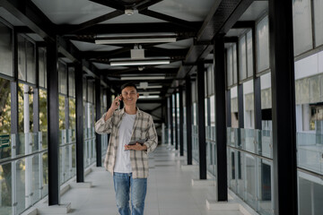 Fototapeta premium Smiling Asian man walking and talking phone with client and holding tablet on his hand in a modern glass hallway. Work from anywhere. Remote work. outdoor.