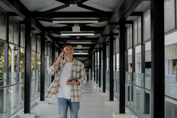 Fototapeta premium happy Asian man walking, looking outside and talking phone with client and holding tablet on his hand in a modern glass hallway. Work from anywhere. Remote work. outdoor.