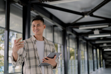 Fototapeta premium Smiling and video call with his friend and holding tablet on his hand in a modern glass hallway. Work from anywhere. Remote work. outdoor.