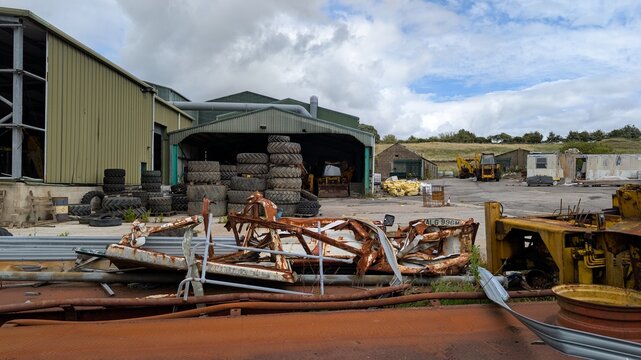 Rusty equipment facility. Deserted scrapyard worksite in rural countryside premises. Industrial tractor tyres piled up. Declining farming agricultural business closures. Abandoned rusting wasteland.