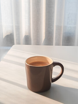 Dark brown coffee cup on a white table by a window with warm morning sunlight. Cozy minimalist lifestyle concept.