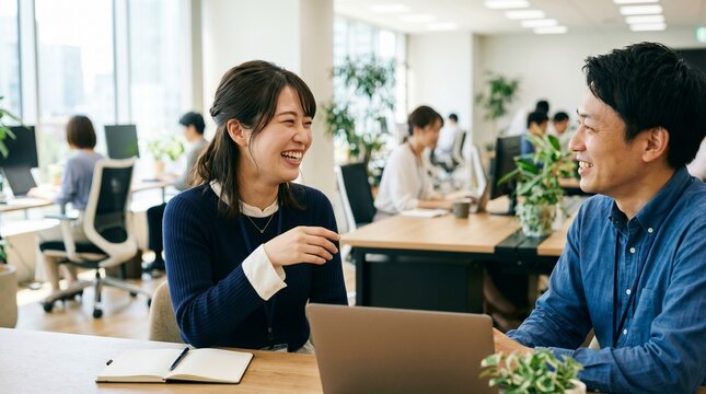良好な職場環境やポジティブなコミュニケーションを象徴する、明るいオフィスで同僚と談笑する日本人女性の新入社員