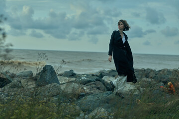 Fototapeta premium Woman walks along a rocky shore in a dark coat, windy weather, distant sea and cloudy sky, creating a moody, cinematic scene of solitude and exploration
